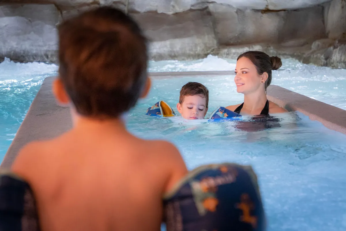 Drei Personen schwimmen in einem Schwimmbecken mit Wasserstrahlen, darunter ein Kind mit Schwimmflügeln.