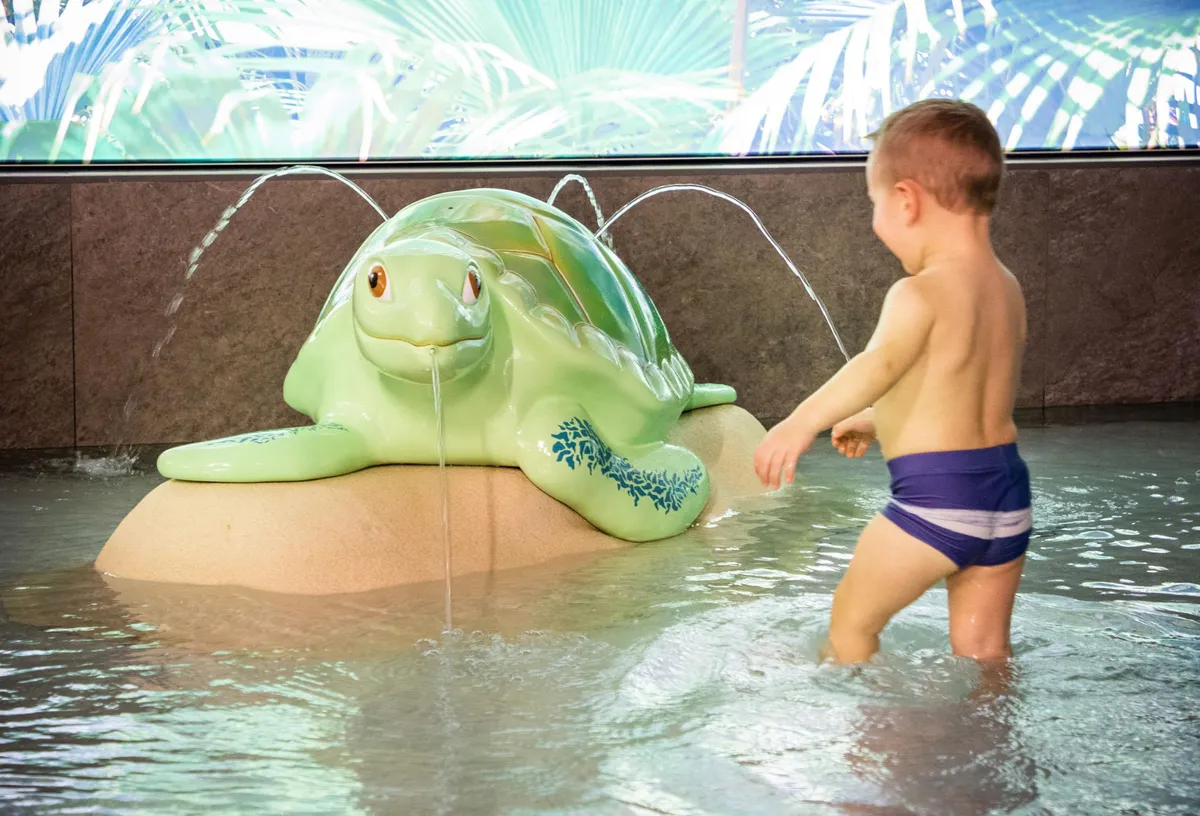 Child in blue swimsuit playing with a turtle sculpture from which water flows into a shallow pool