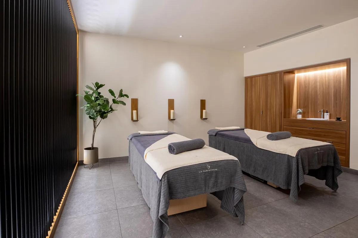 Zwei Massagetische mit grauen und weißen Handtüchern, eine grüne Pflanze und Wandlampen in einem Raum mit beigefarbenen Wänden und grauem Boden