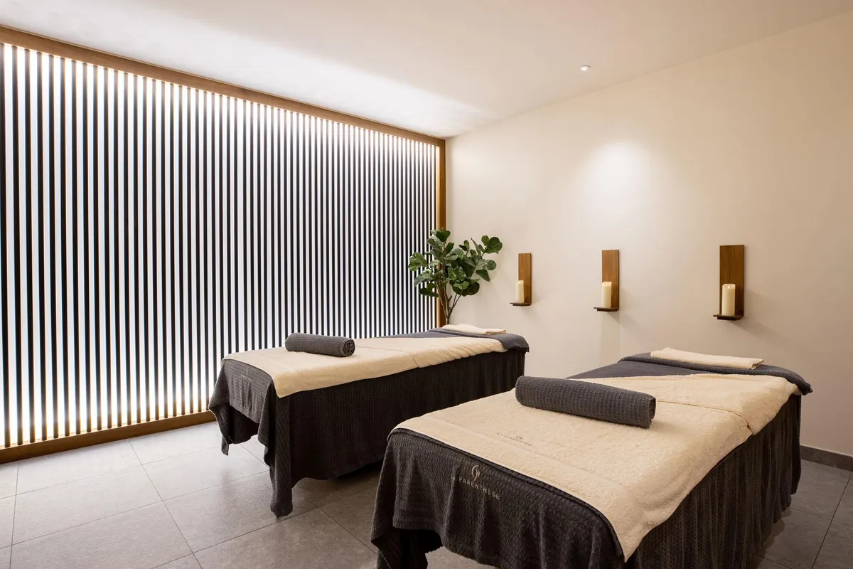 Two massage tables with towels, lit by wall lamps, in a room with beige walls and a slatted window.