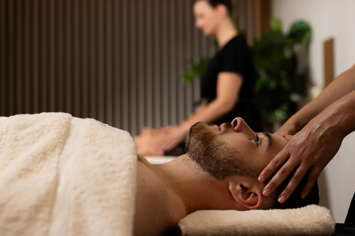 A man lying on his stomach receives a neck massage from a professional, in an indoor setting with plants in the background.