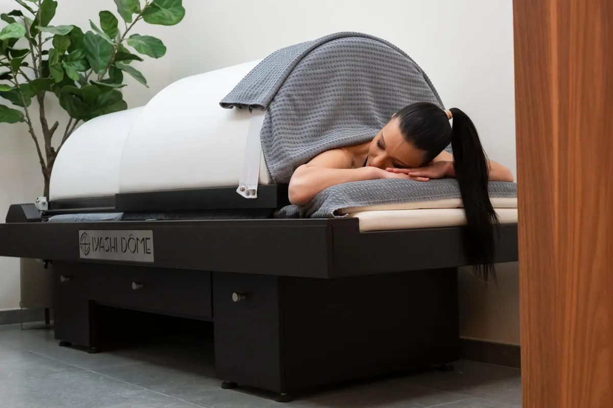 A person lying on a massage bed covered with a gray blanket in a room with a green plant.
