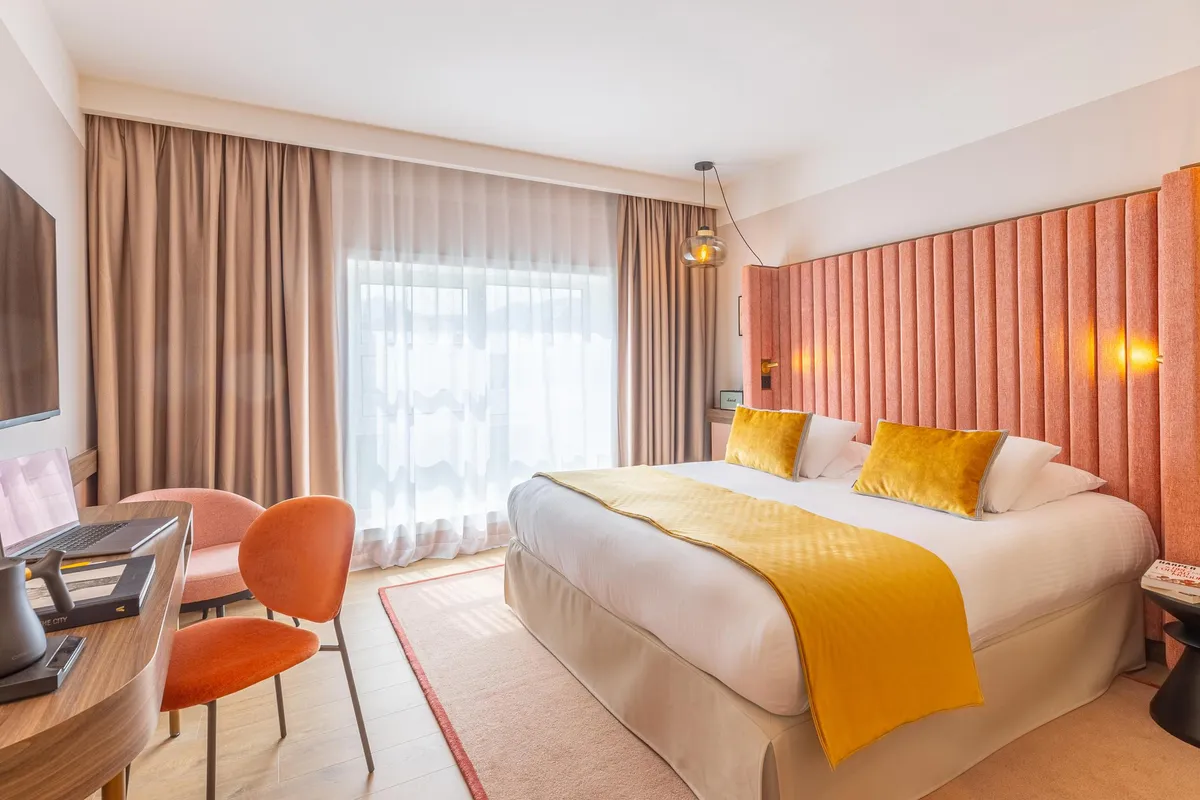 Hotelzimmer mit einem Doppelbett, zwei Stühlen und einem Schreibtisch neben einem Fenster mit Vorhängen