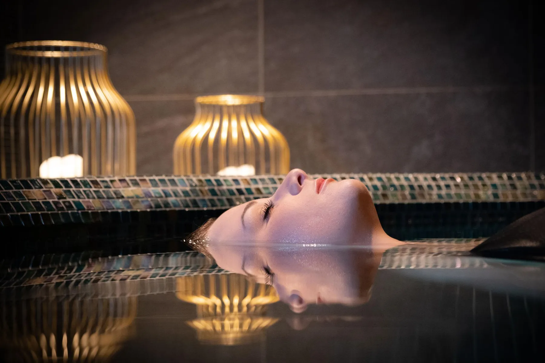 A person is lying in a pool of water, their head above the water, with lamps in the background.