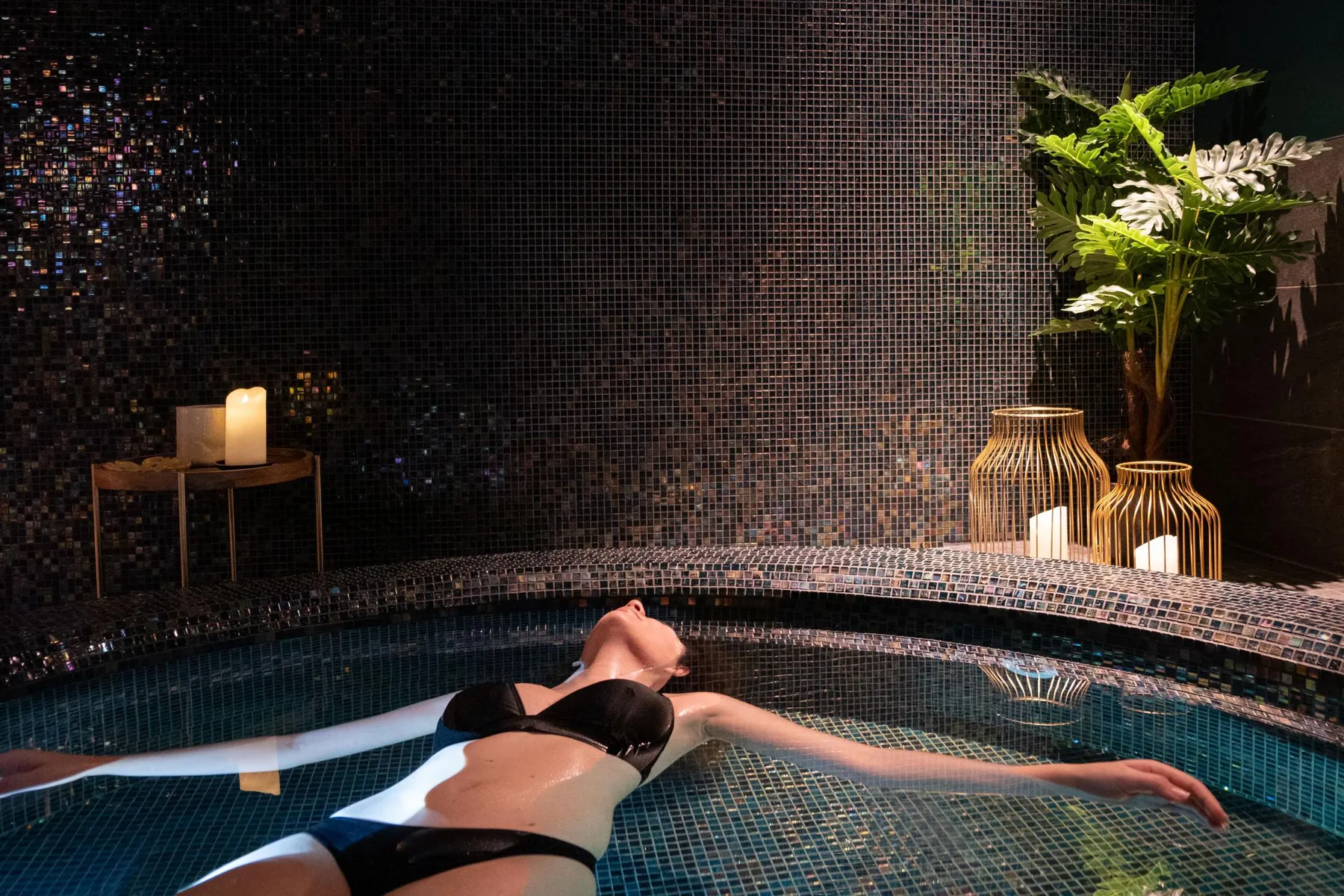 A person in a black swimsuit floats on their back in an indoor pool with a mosaic wall and illuminated decorations.