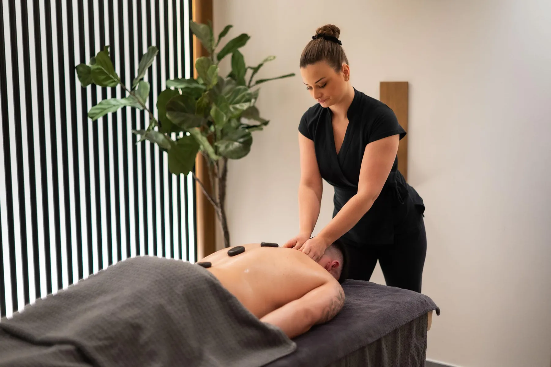 A woman in black attire is performing a massage on the back of a person lying on a table covered with a towel, with a green plant nearby.