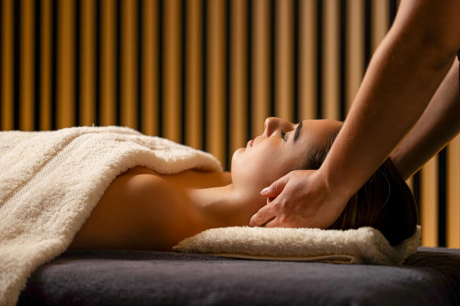 A person receives a massage on a massage bed covered with towels, in a room with vertically striped walls.