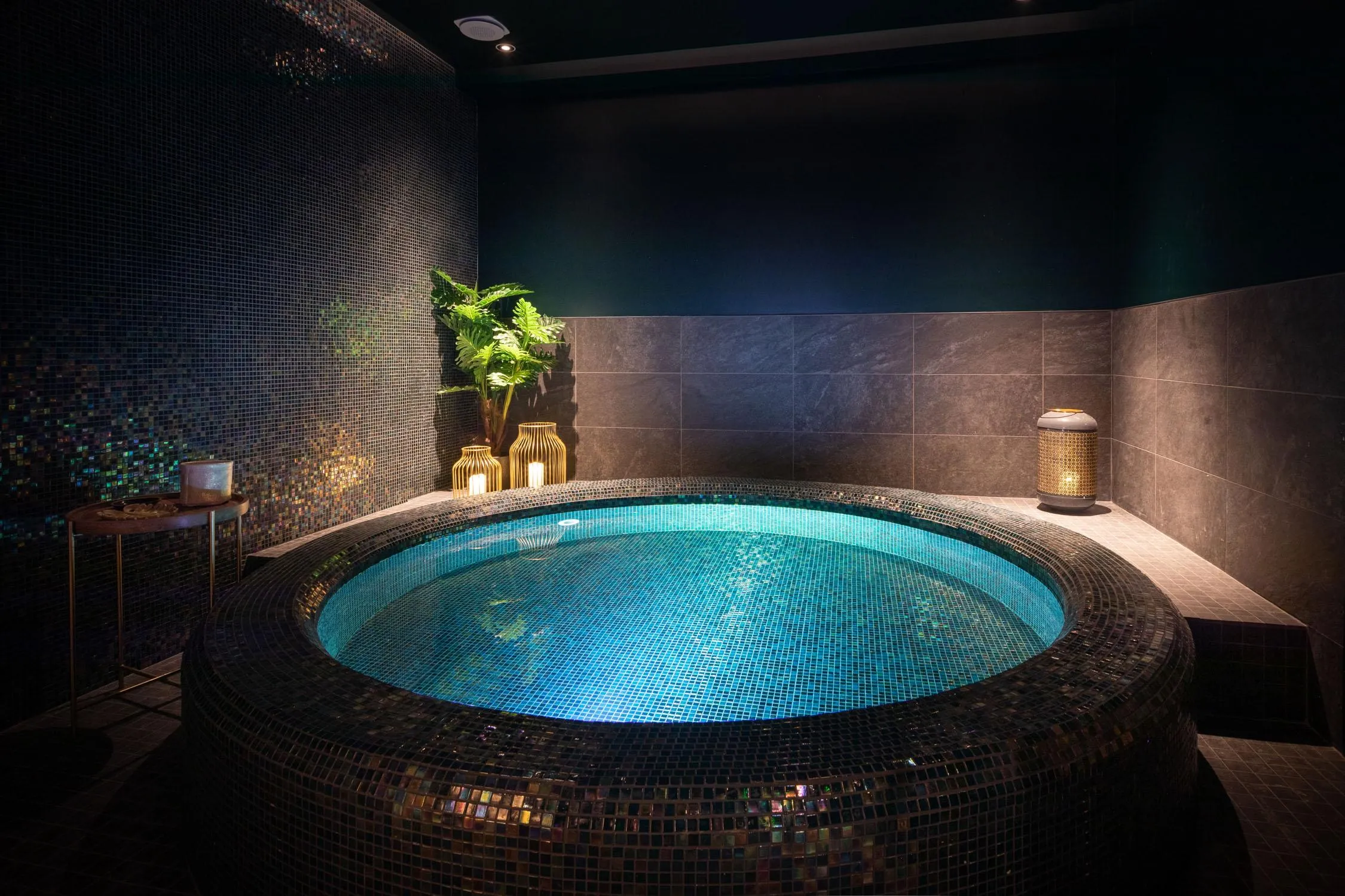 Runder Jacuzzi mit blauer Beleuchtung, umgeben von Mosaiken, in einem Raum mit dunklen Wänden, Pflanzen und Laternen
