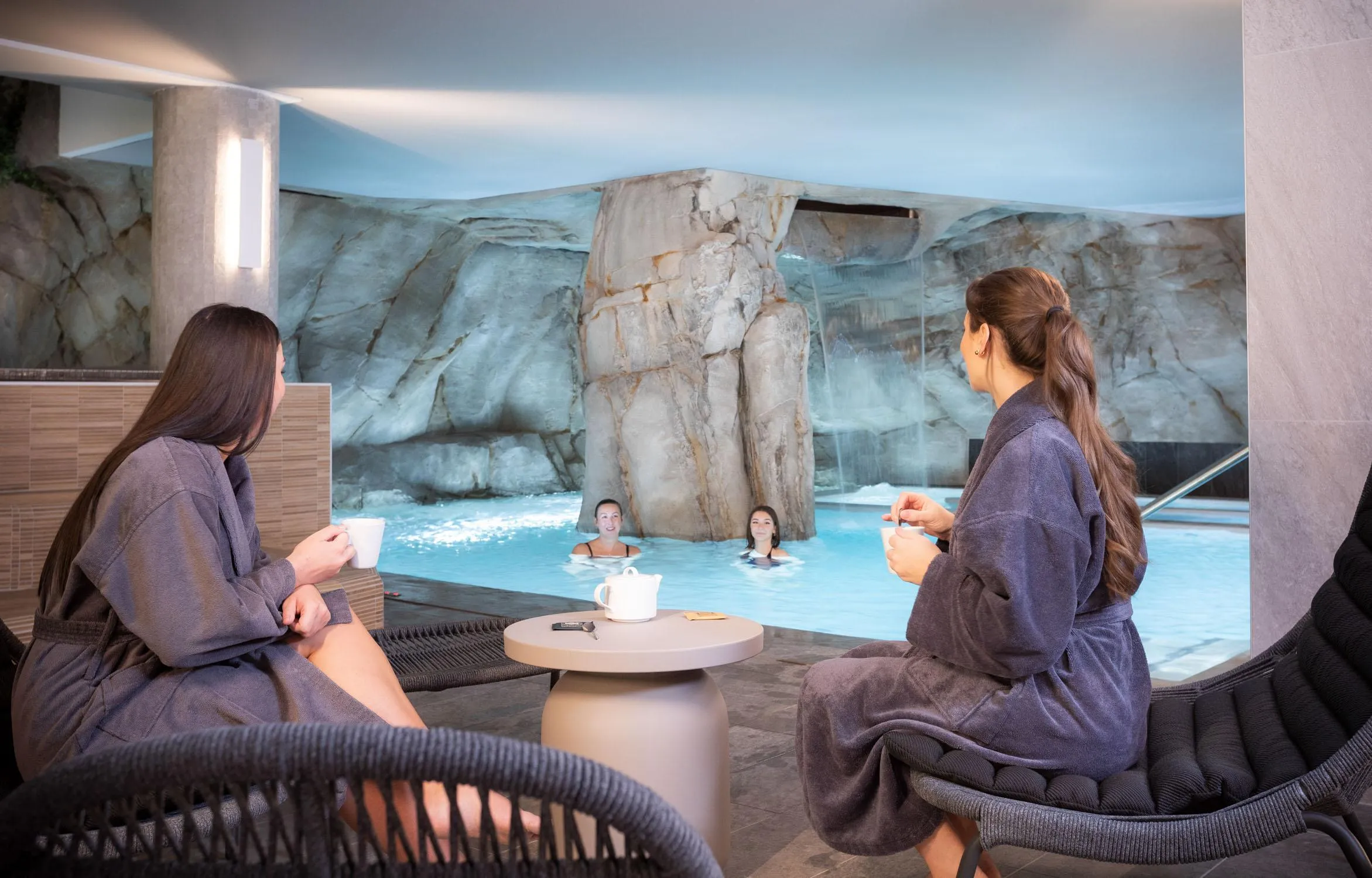 Two people sitting in robes talk near an indoor pool with an artificial waterfall, while two others swim in the water.