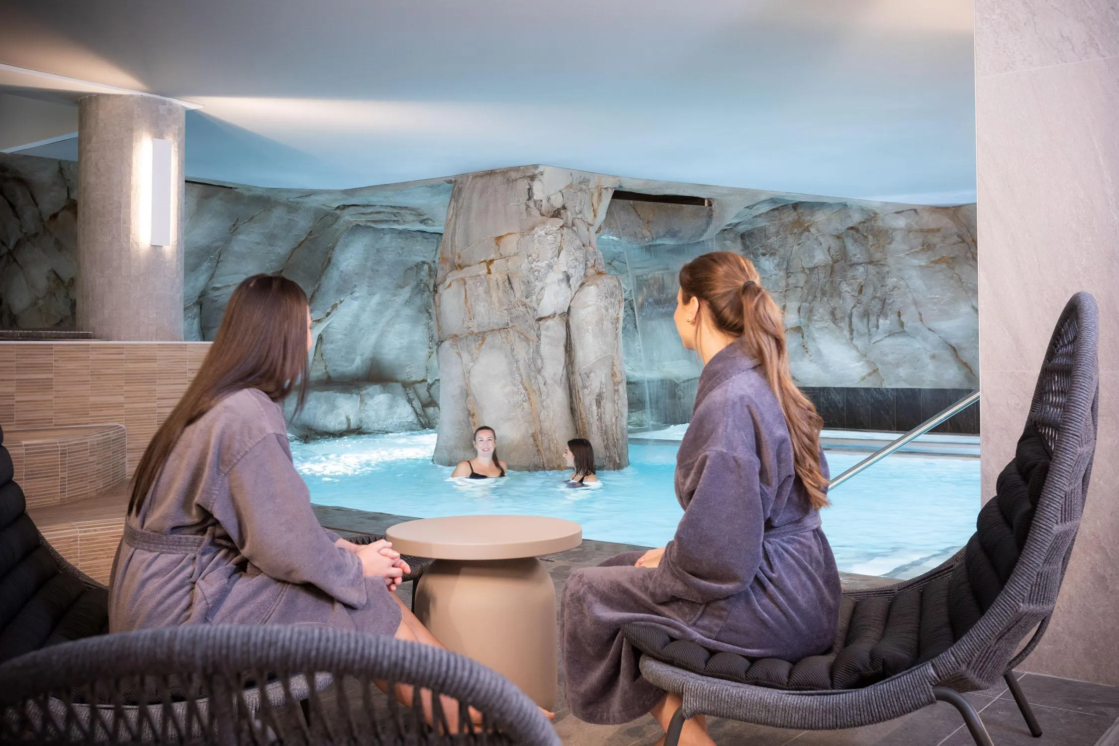 Zwei Personen in Badekleidung sitzen und beobachten ein Wasserbecken mit Felsen und Wasserfällen, in dem zwei weitere Personen schwimmen.