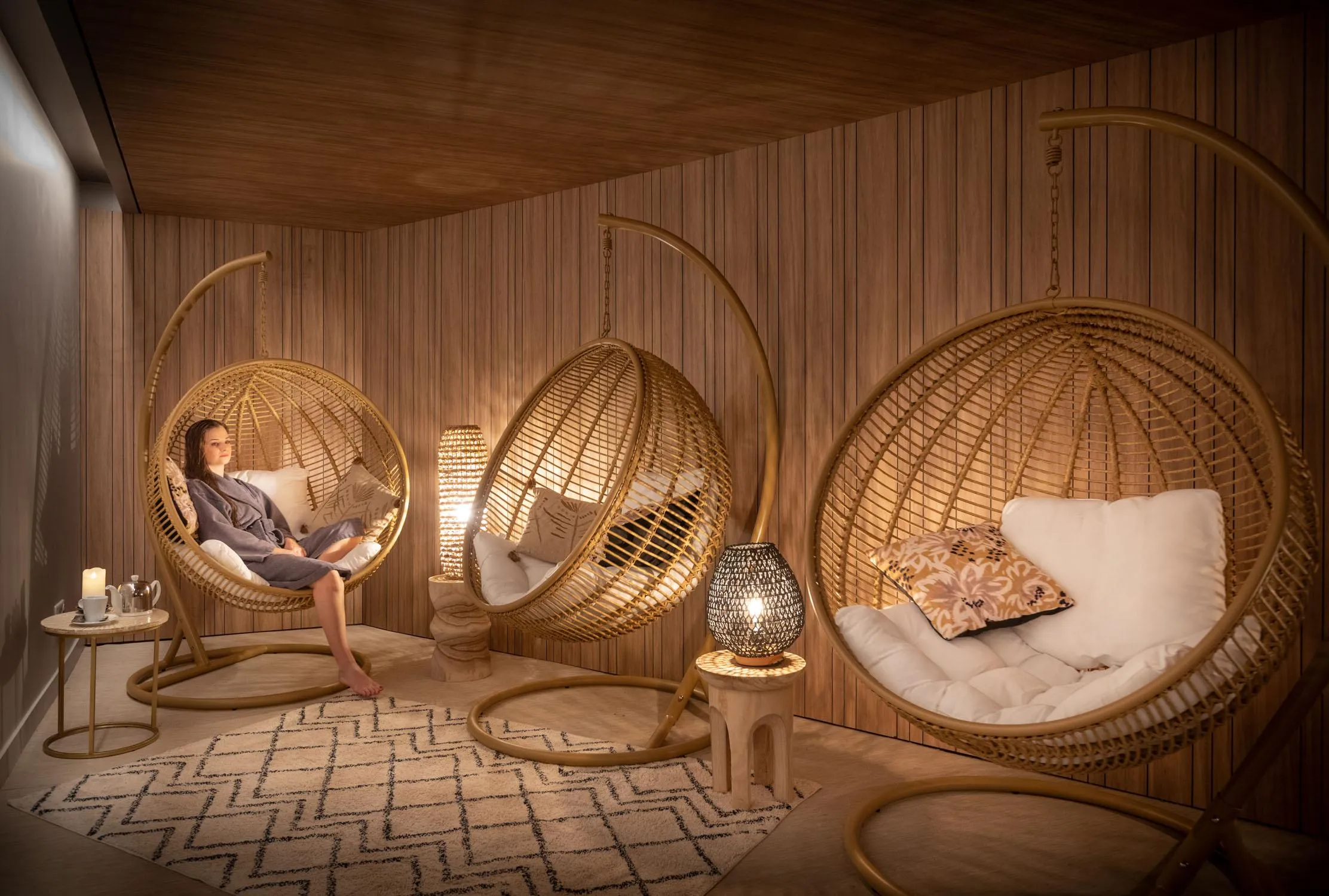 Three people sitting in hanging wicker chairs in a room with wooden walls, lamps, and cushions.