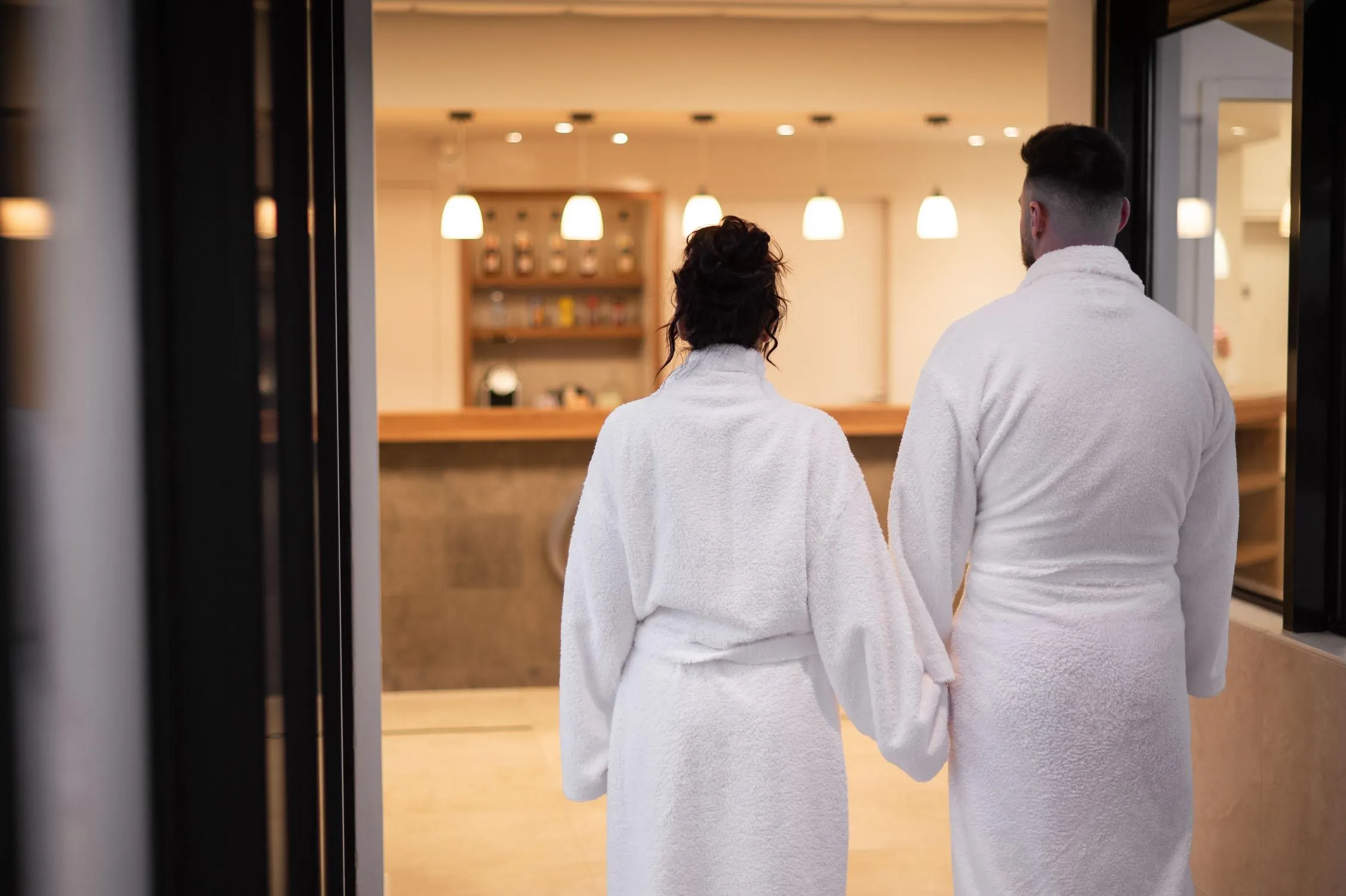 A couple in white robes stands with their back to the camera in front of a spa display, hand in hand, observing the lit interior.