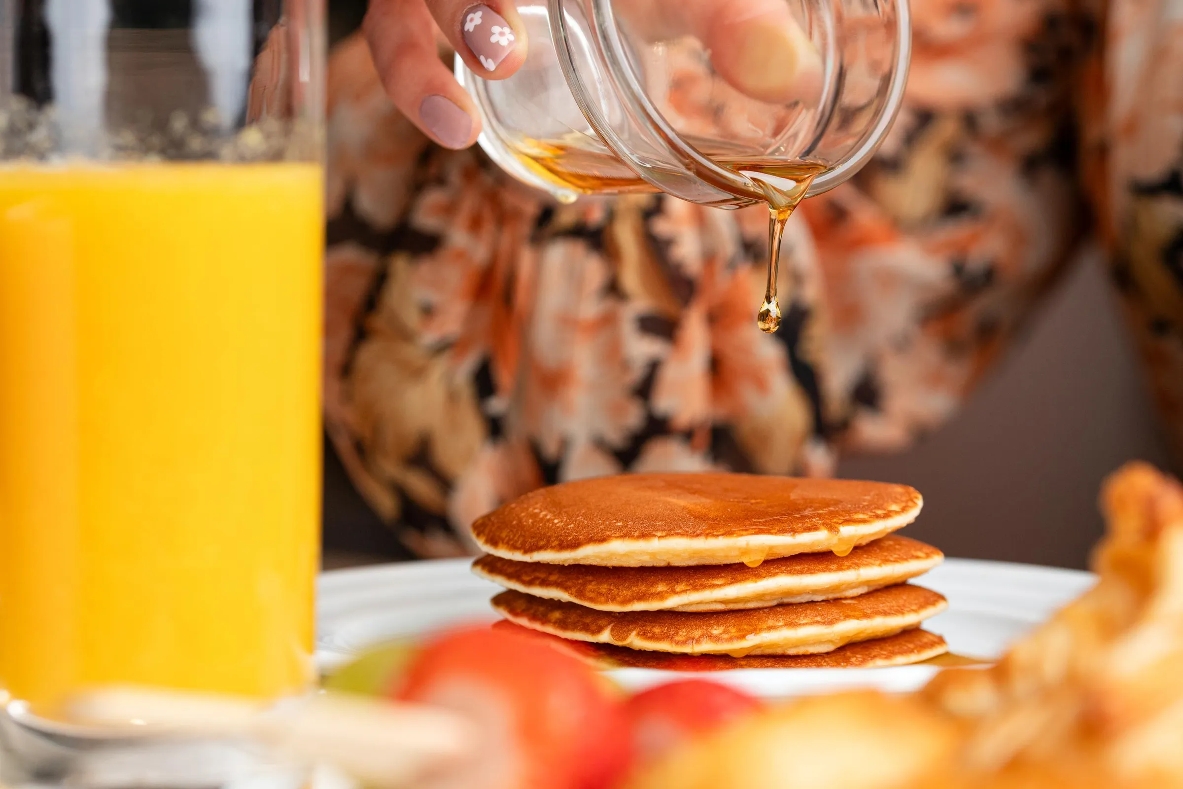 A hand pours syrup over a stack of pancakes, with a glass of orange juice to the left.