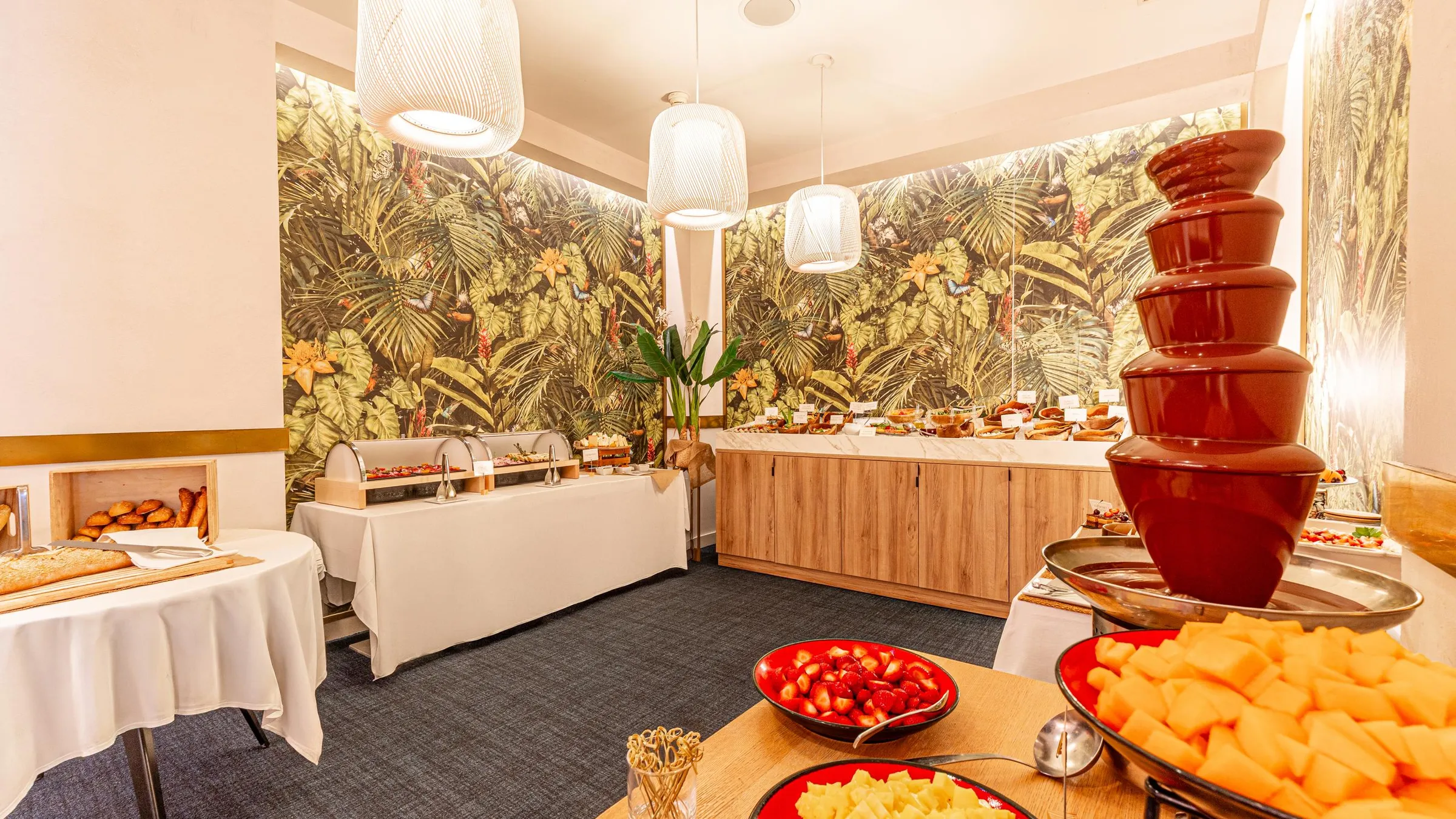 Buffet mit verschiedenen Gerichten, darunter eine Schokoladenbrunnen, in einem Raum mit tropischen Mustern und beleuchtet durch Hängelampen
