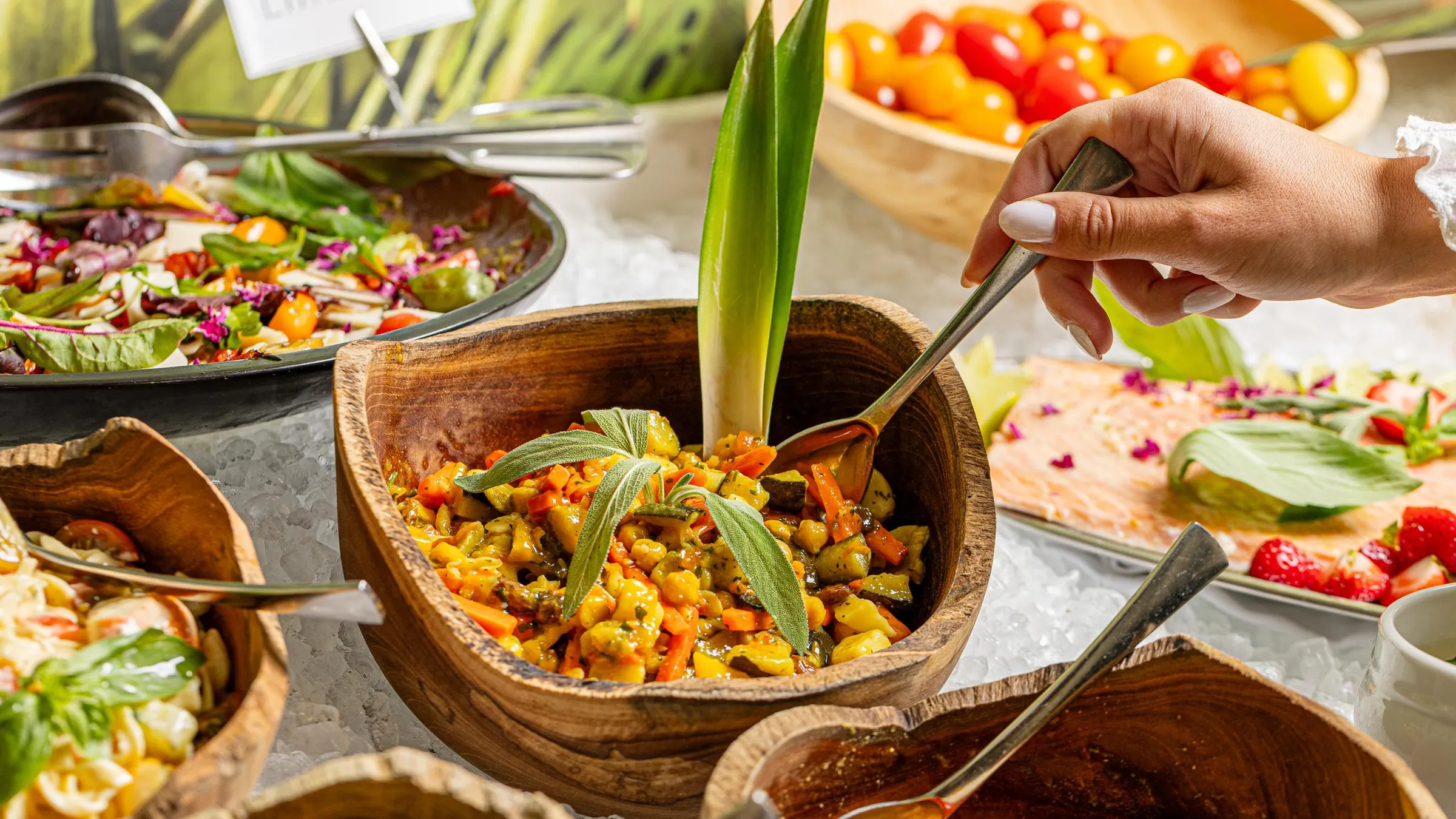 Eine Hand serviert eine Gemüsezubereitung in einer Holzschüssel auf einem Buffet mit verschiedenen Gerichten.