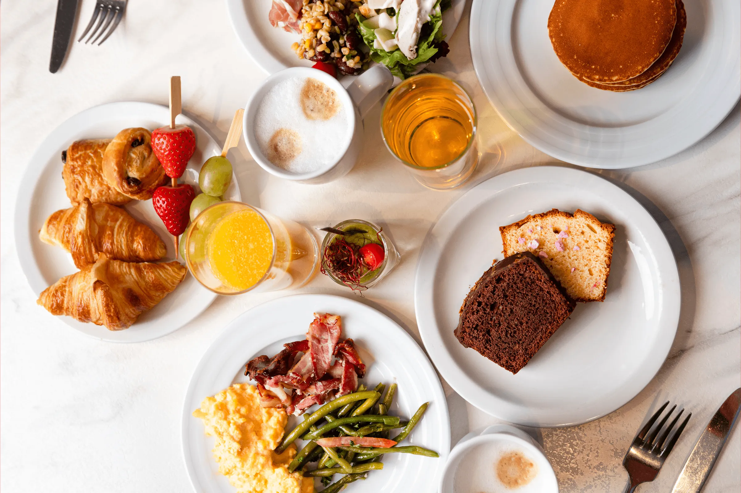 An assortment of dishes and drinks arranged on a table, including croissants, fruits, vegetables, eggs, bread, and hot and cold beverages.
