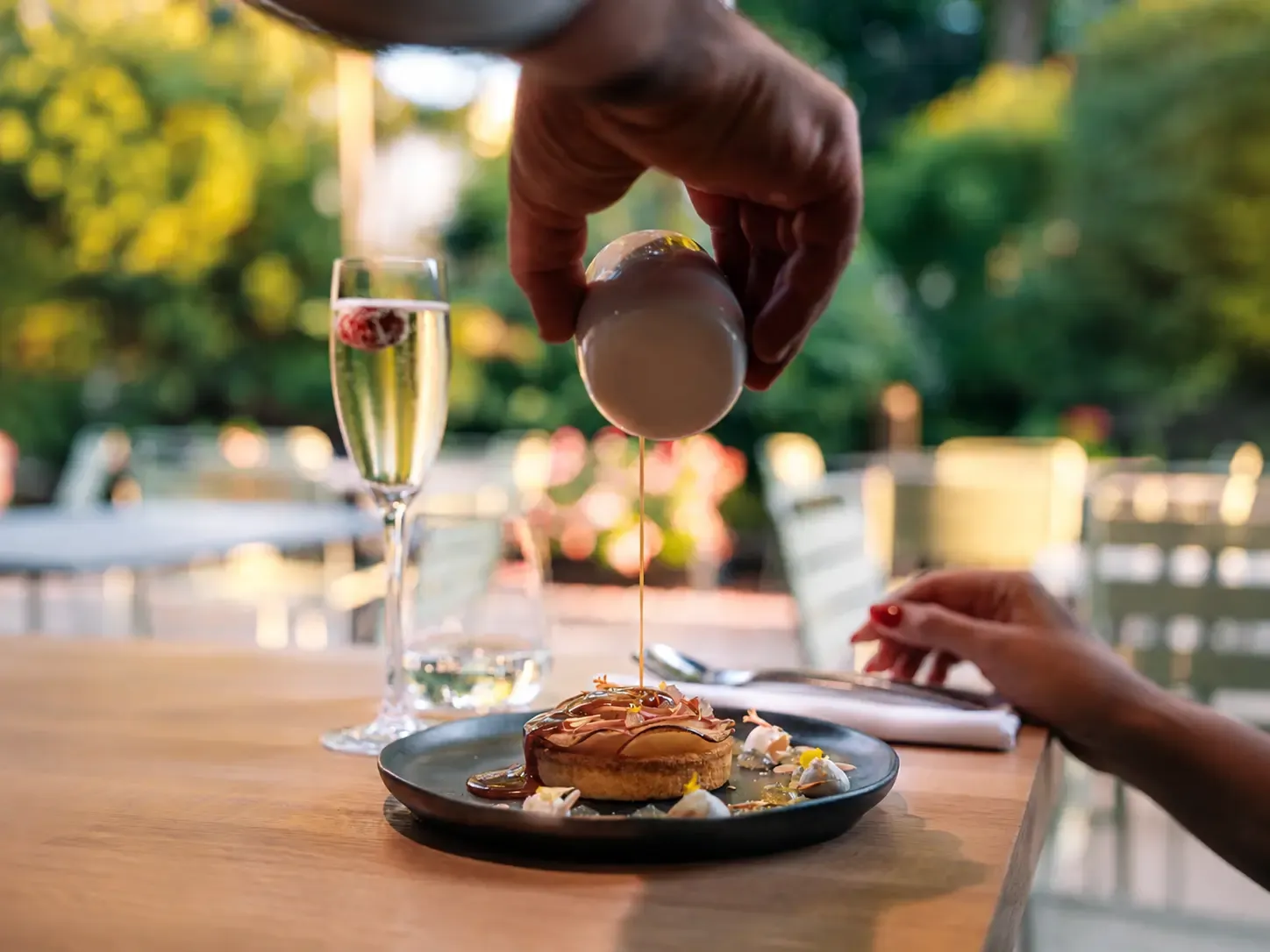 Une main verse un filet de sauce sur un dessert placé sur une assiette noire, avec un verre de champagne à proximité