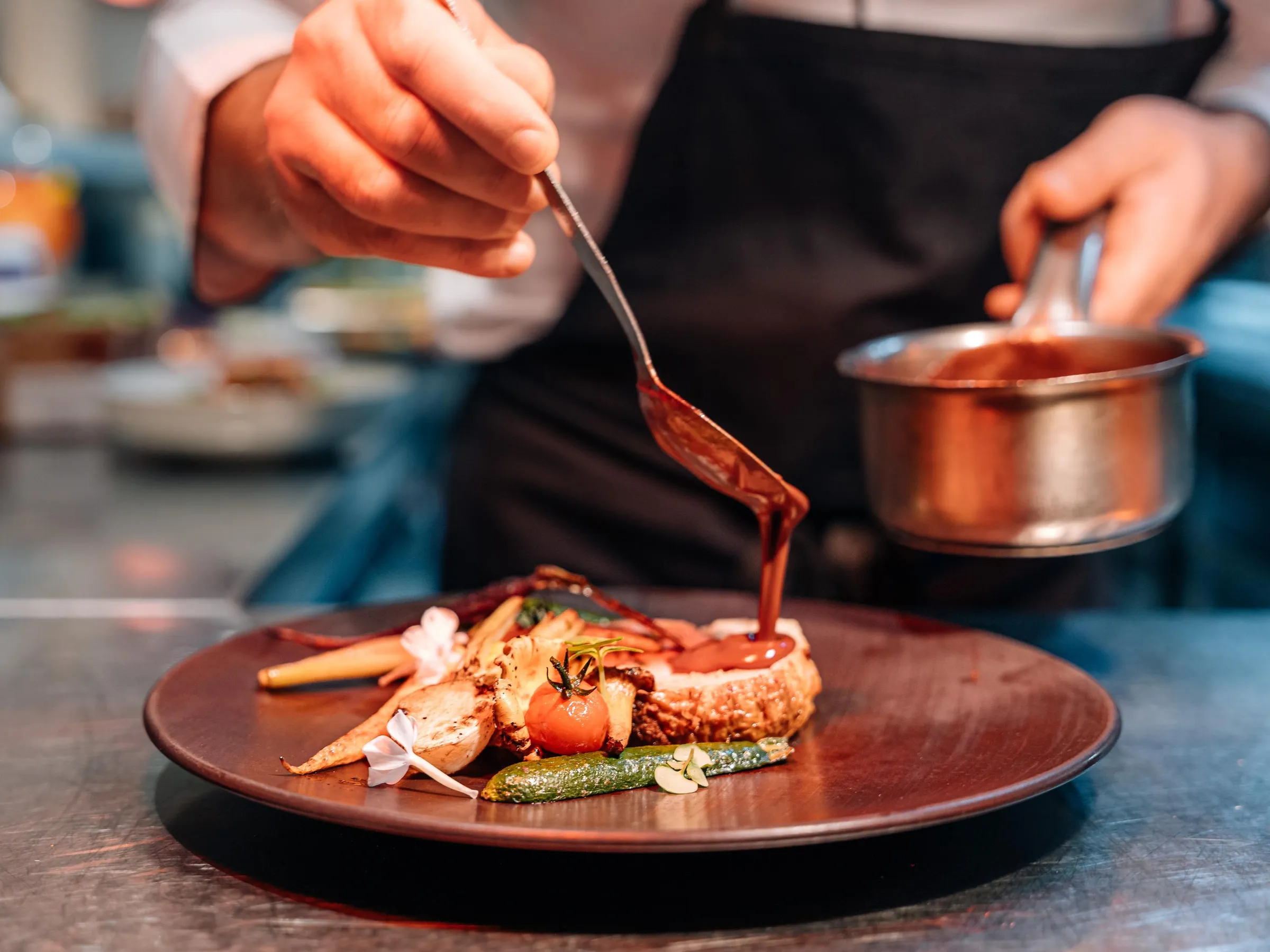 A chef cooks a sauce over a dish composed of vegetables and grilled meat.