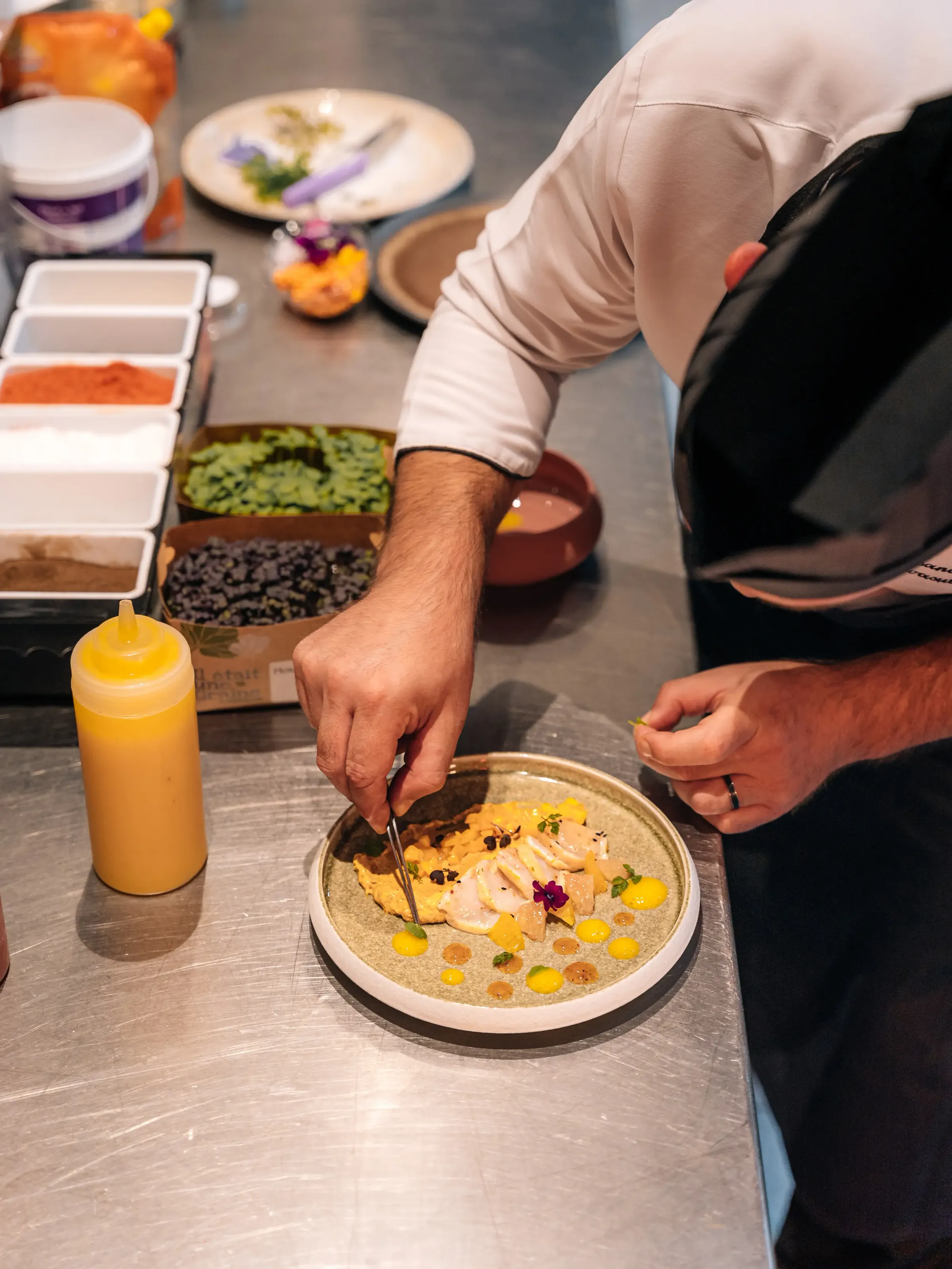 Un chef en veste blanche prépare une assiette de poisson garnie de légumes et de sauces colorées sur un plan de travail de cuisine