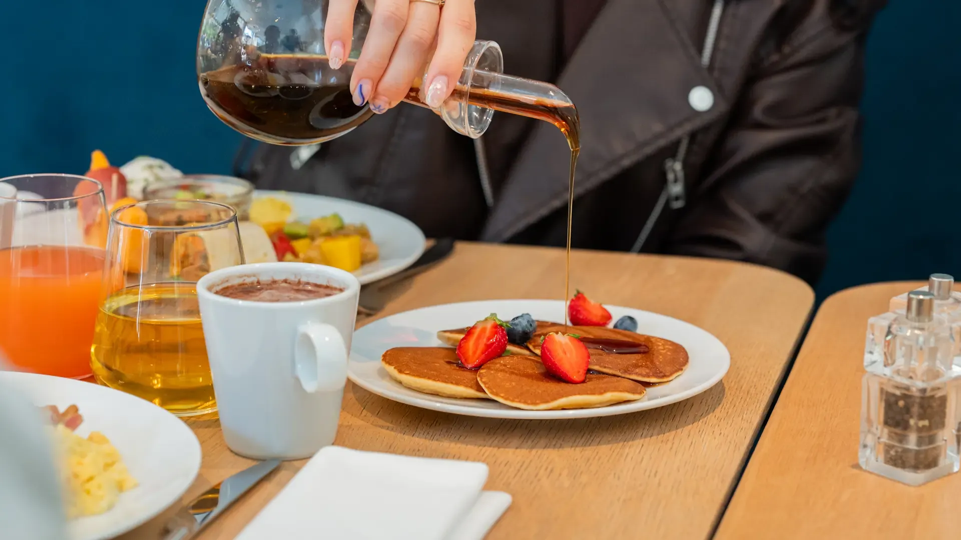 Eine Person gießt Sirup über mit Früchten belegte Pfannkuchen, die mit Getränken und einer Obstplatte auf einem Tisch serviert werden.