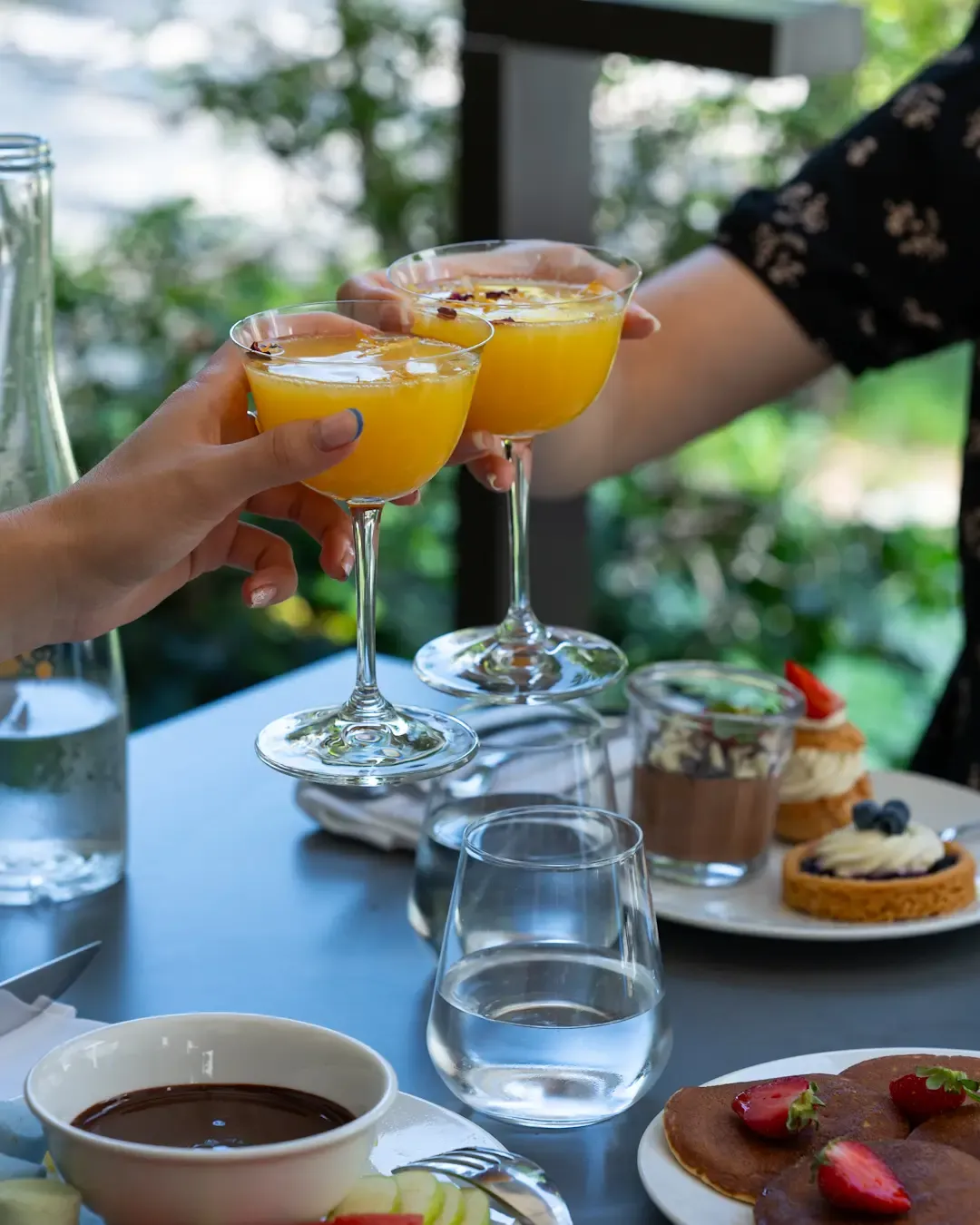 Zwei Hände heben orangerote Cocktailgläser mit Zitruszesten hoch, um einen Tisch mit Desserts und Getränken.