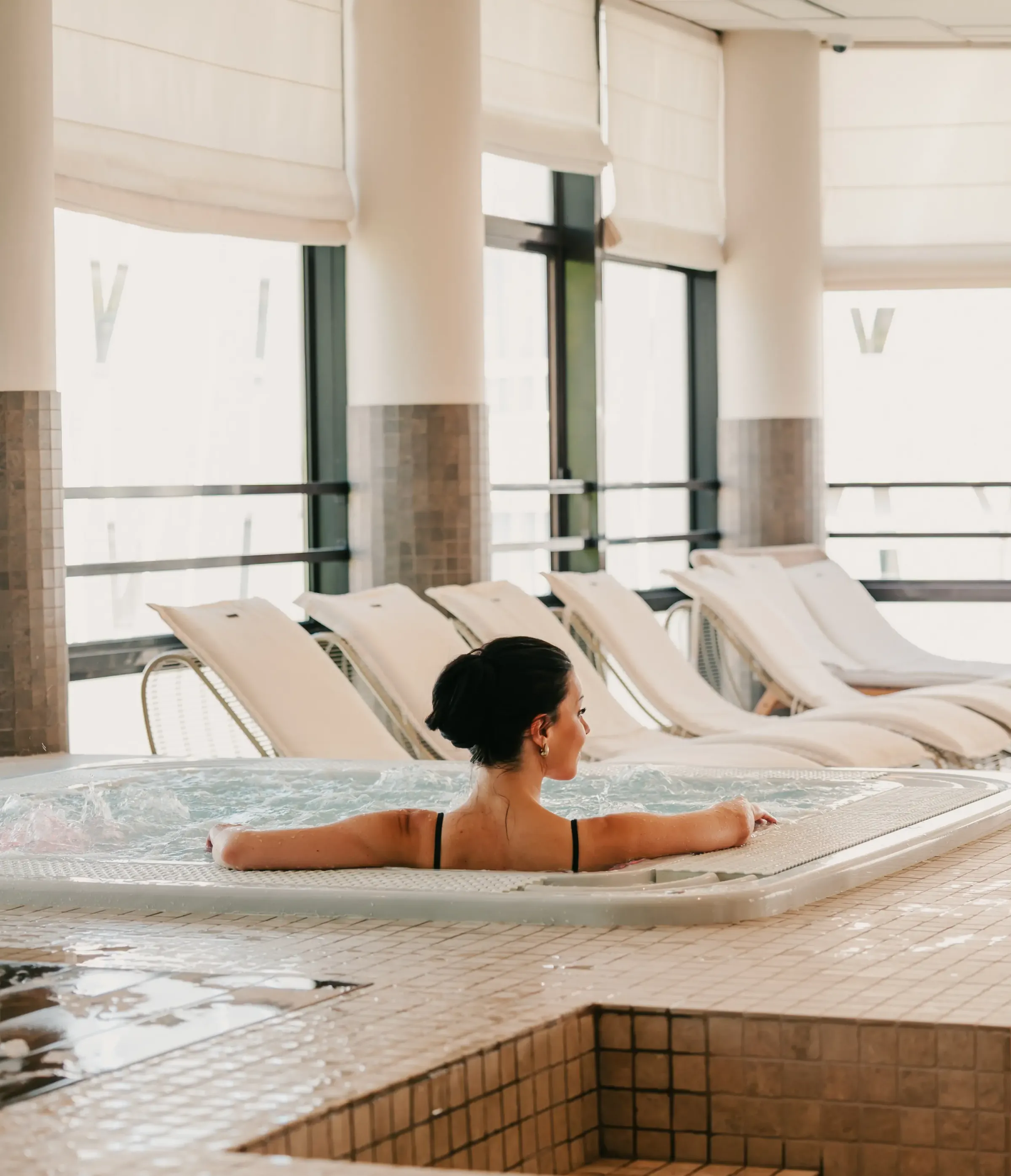 Eine Frau in einem schwarzen Bikini liegt in einem Whirlpool, umgeben von Liegestühlen nahe großen Fenstern.