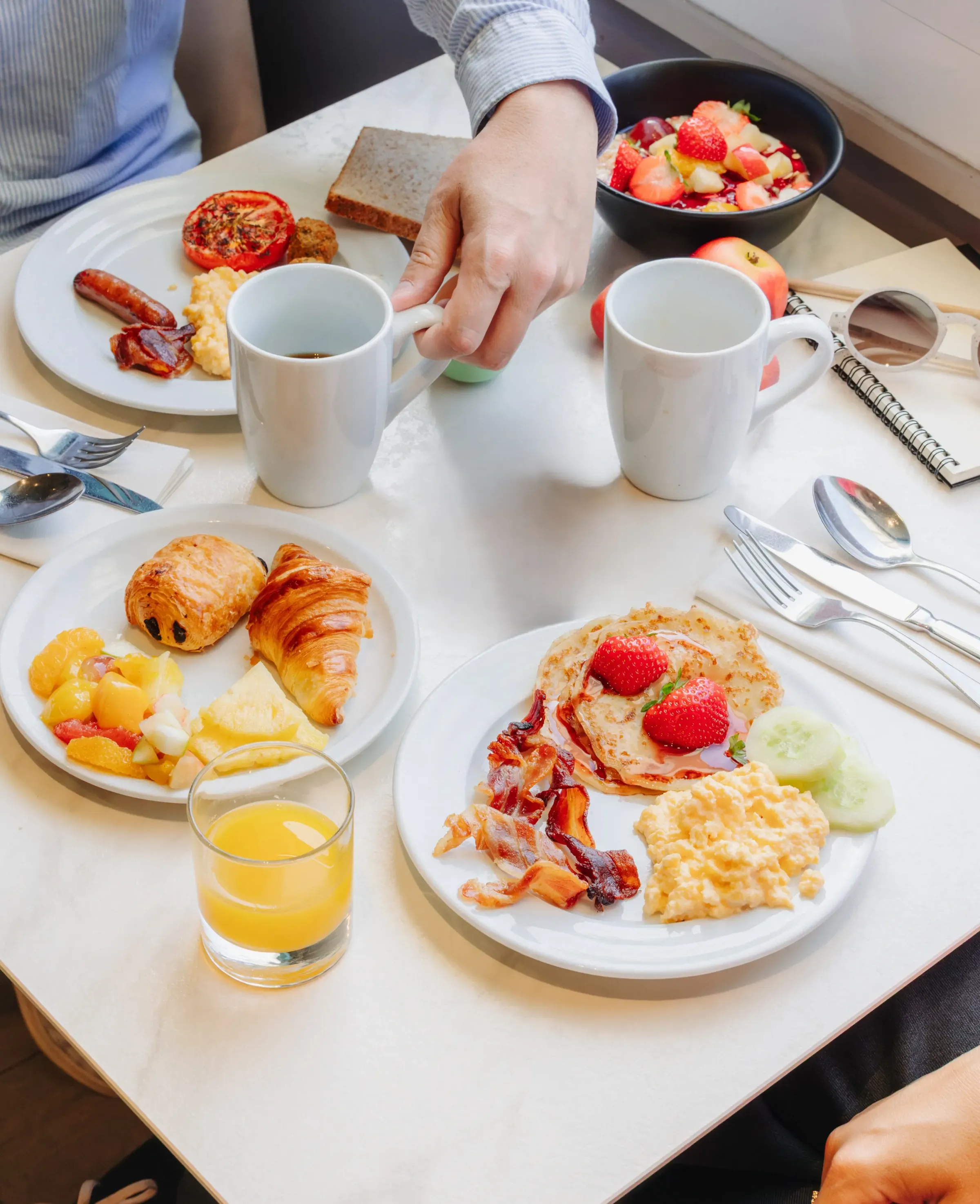 Une table avec des assiettes de petit-déjeuner, des fruits, des boissons et des couverts, une personne tenant une tasse