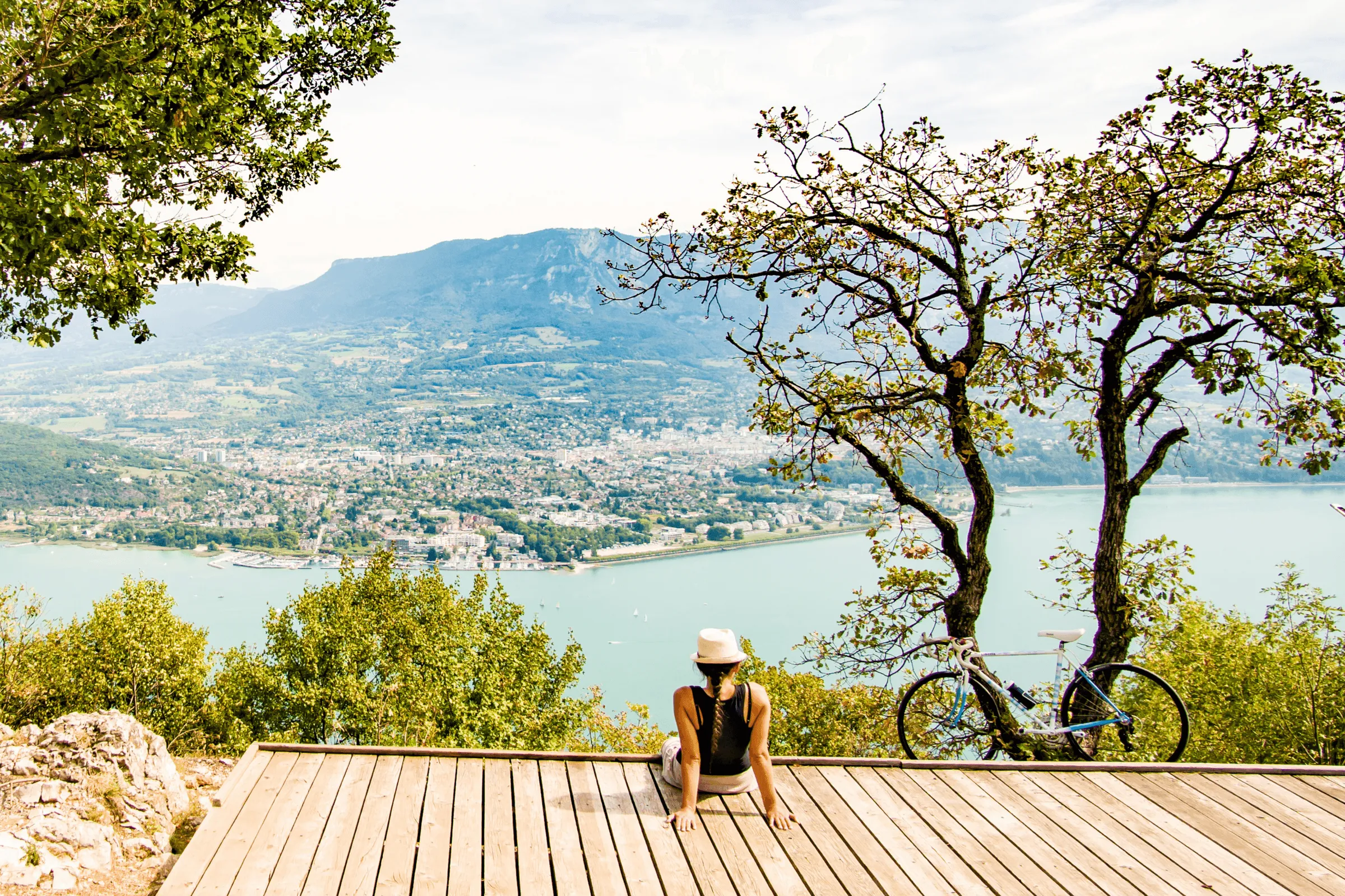 A person sitting on a wooden platform with a bicycle beside them, gazing at a lake and a city surrounded by mountains.