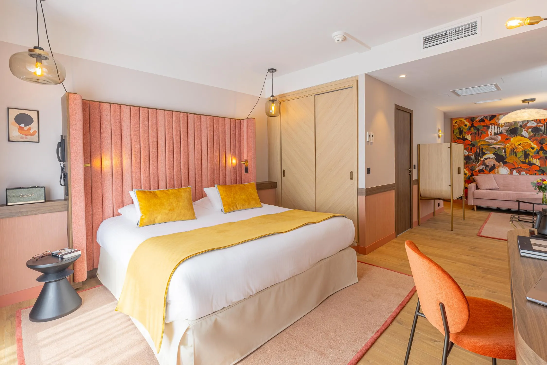 Hotelzimmer mit einem Doppelbett mit weißen Laken und einer gelben Decke, einem rosa Kopfteil, einem Nachttisch mit Gegenständen darauf, einer Hängelampe, einem orangen Sessel und einer offenen Tür zu einem Wohnzimmer mit einem Sofa und einem Wandbild