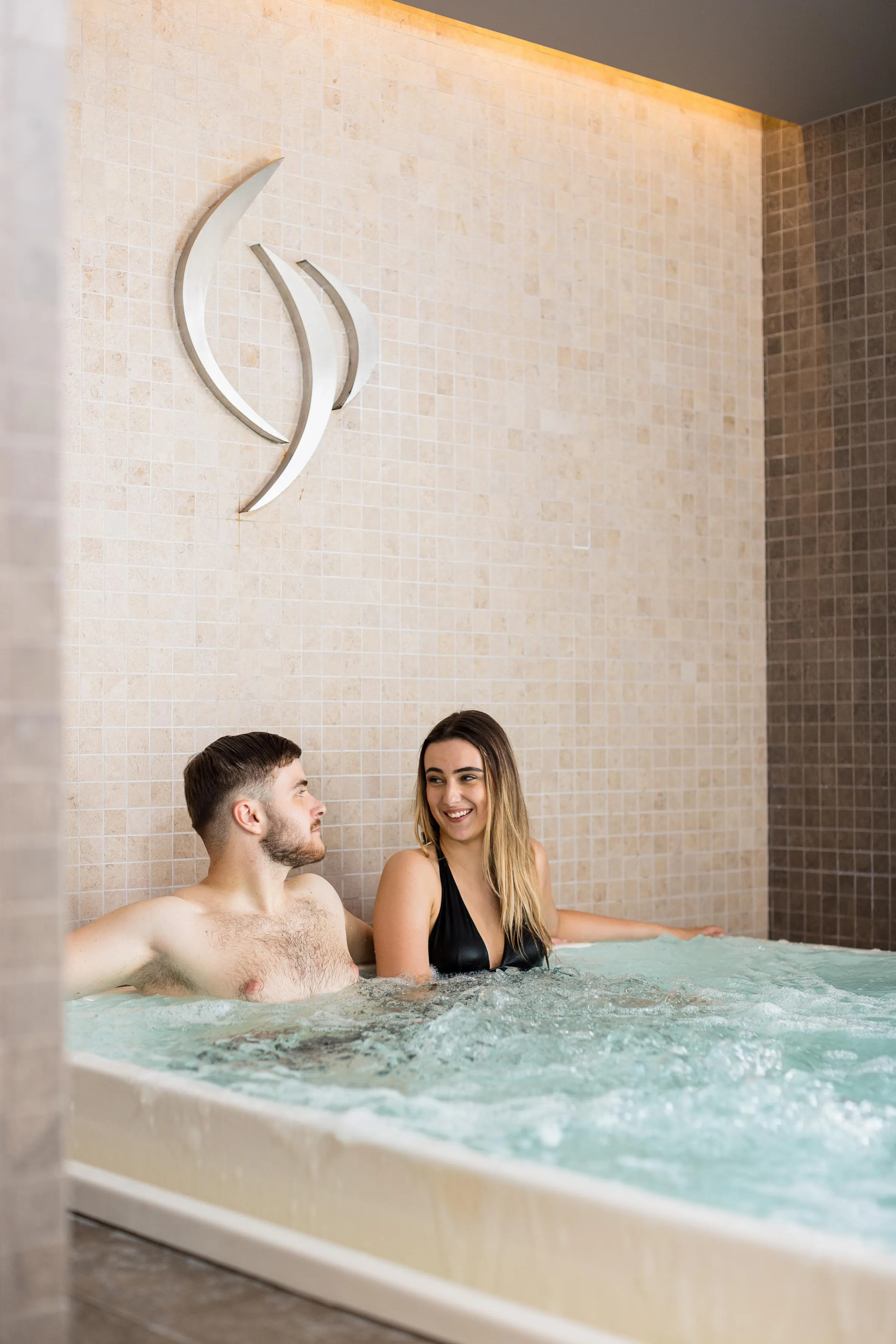 A man and a woman smile in a hot tub with a moon symbol above the tiled wall.