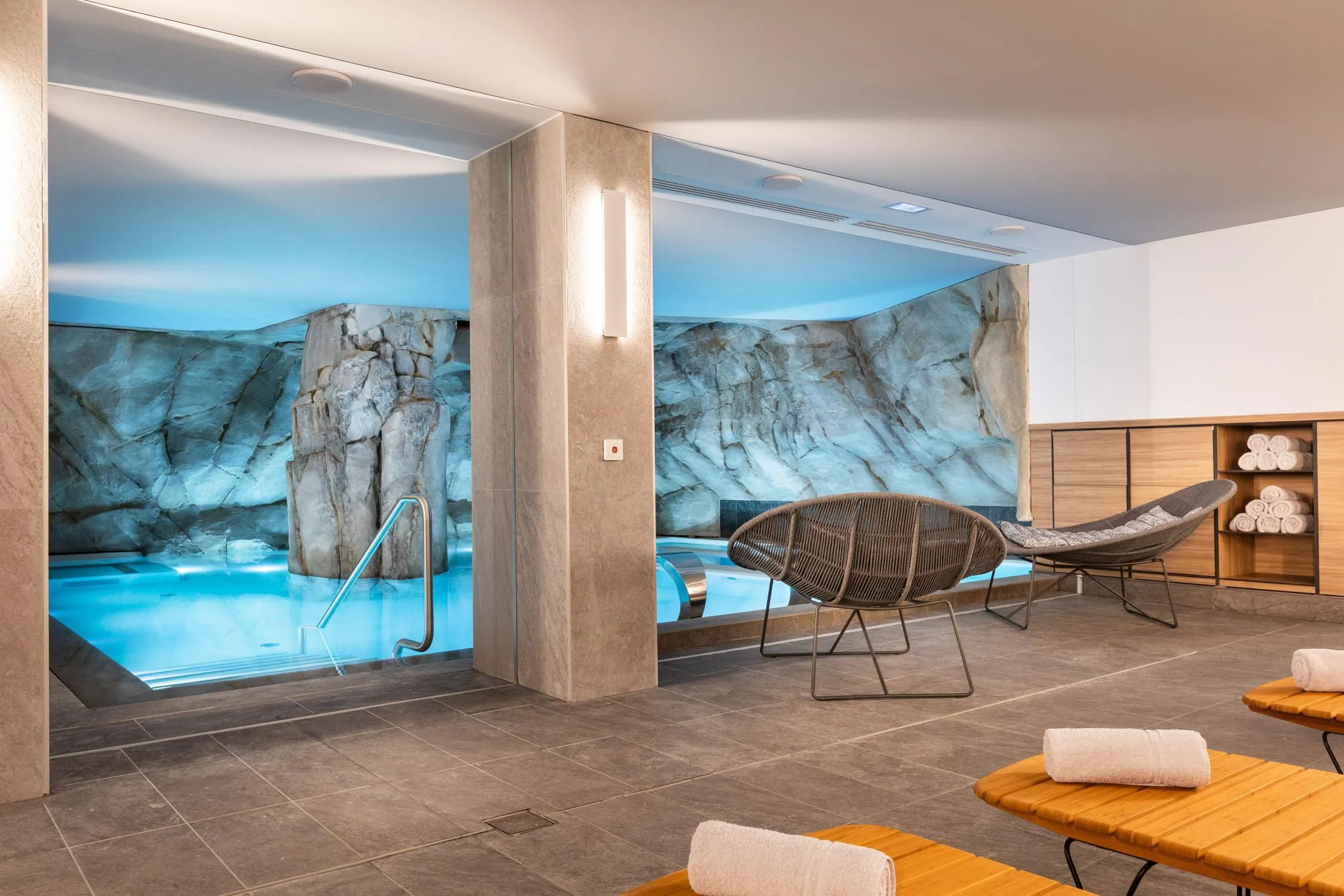 Relaxation room with sun loungers, towels, and a view of a pool through a large glass wall decorated with rocks.