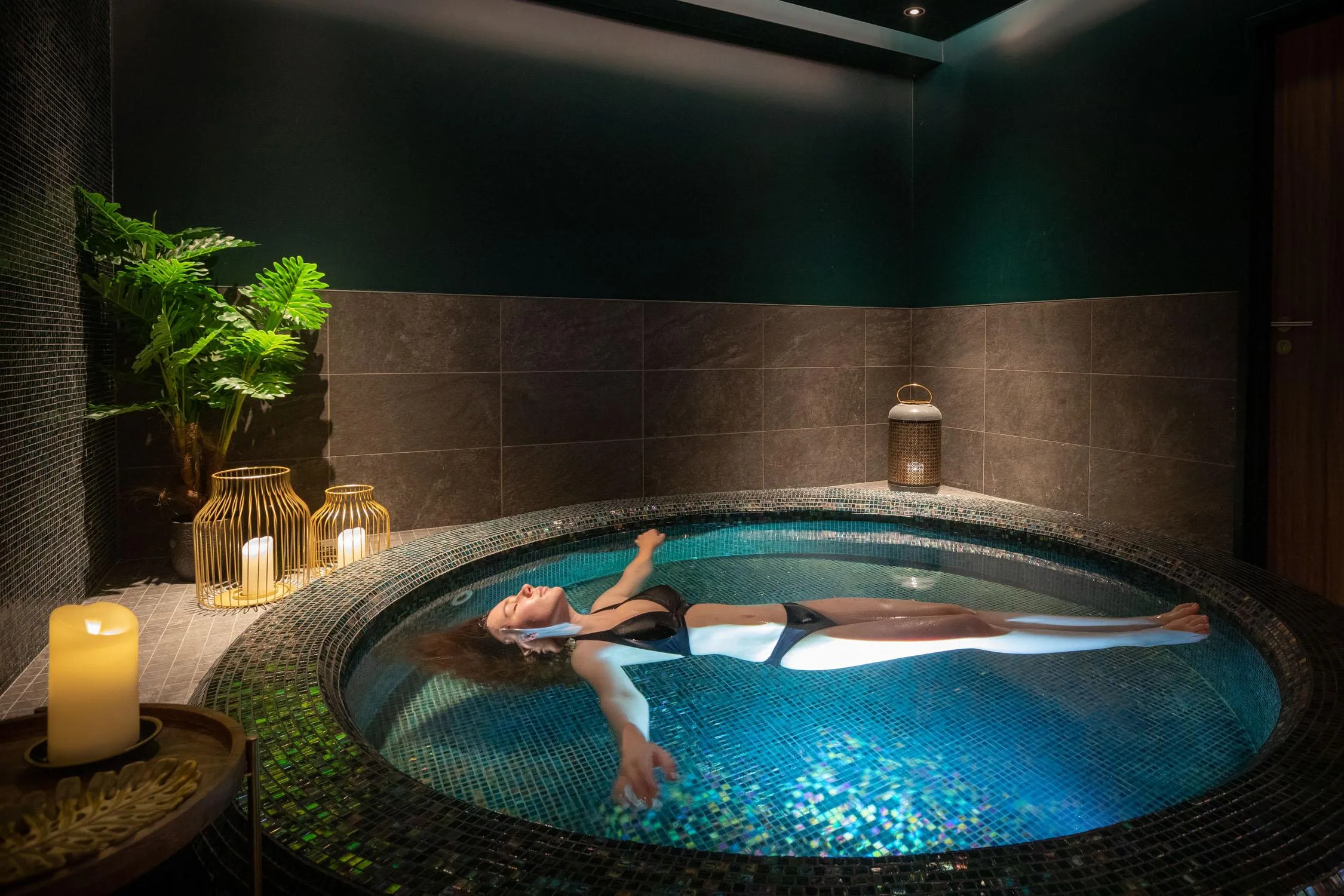 A person lying in a circular hot tub surrounded by candles and green plants.