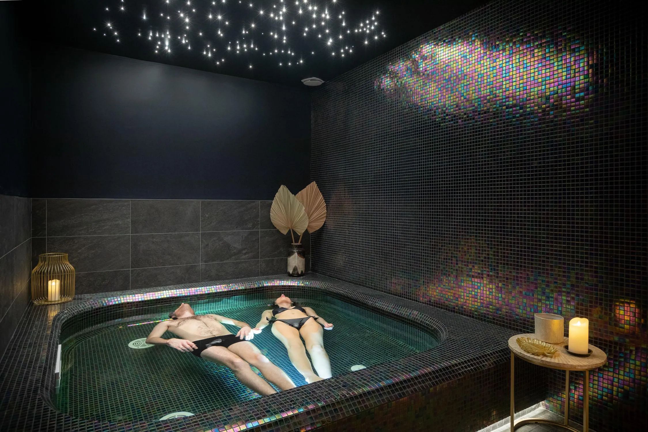 Two people lying in a hot tub, surrounded by colorful mosaics and candles, with a starry ceiling.