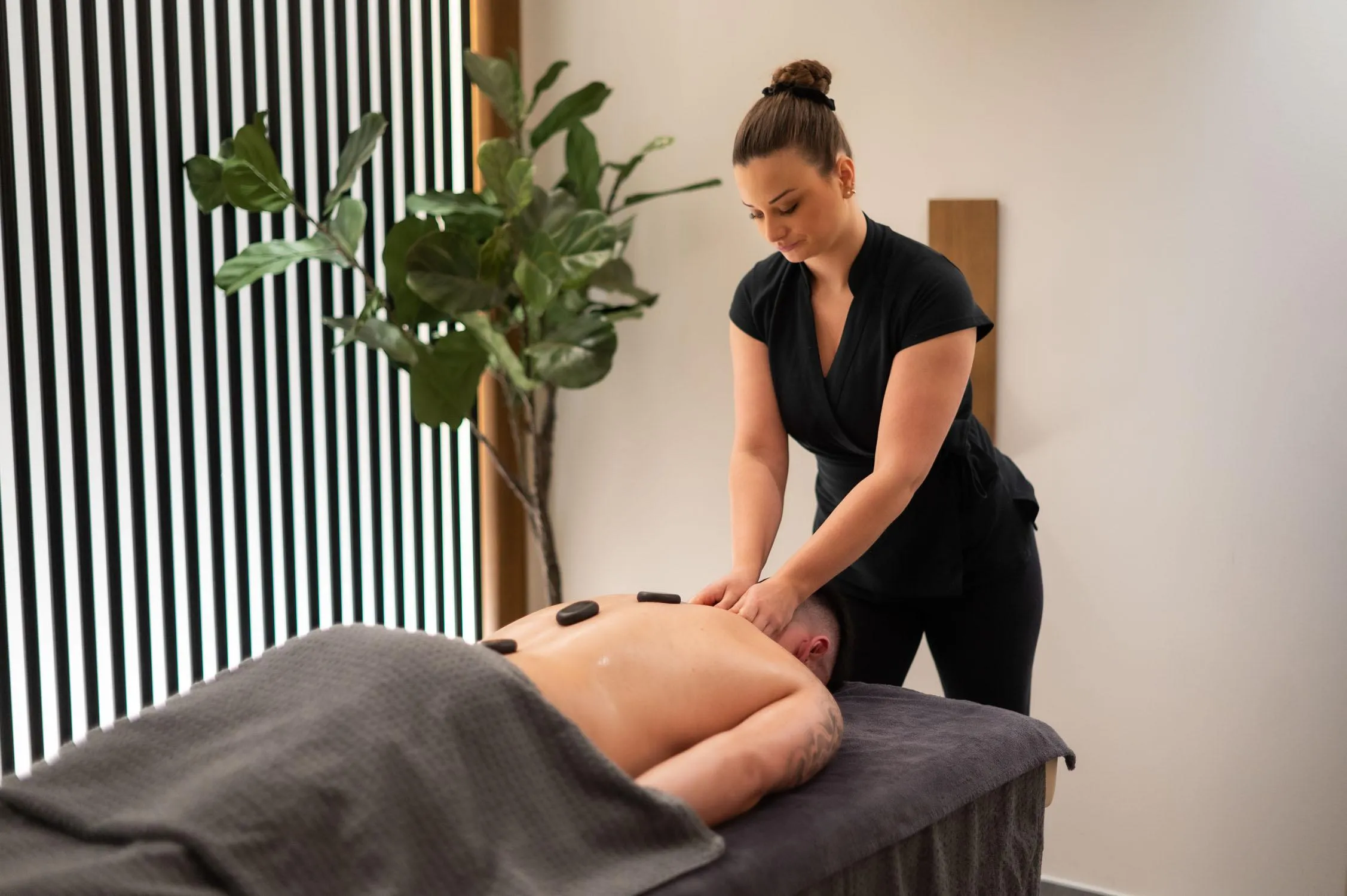 A woman in black attire is performing a massage on the back of a person lying on a table covered with a towel, with a green plant nearby.